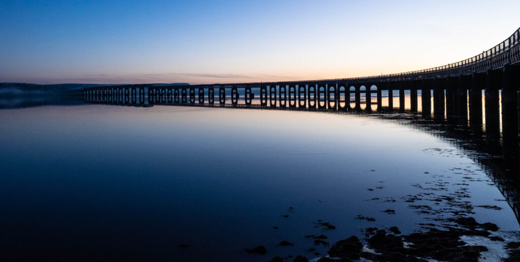 A long windy bridge construction with huge arches. The bridge is over a big lake with a clear blue and pink sky.