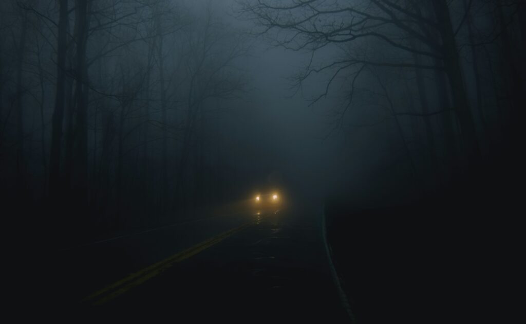 A dark, foggy road with trees surrounding and a singular car driving down it with its headlights on.