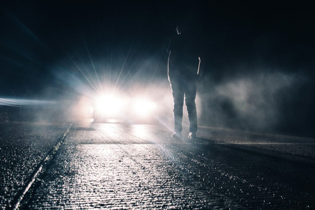 A foggy dark road showing a car stopped with headlights on an a human figure standing in the light.