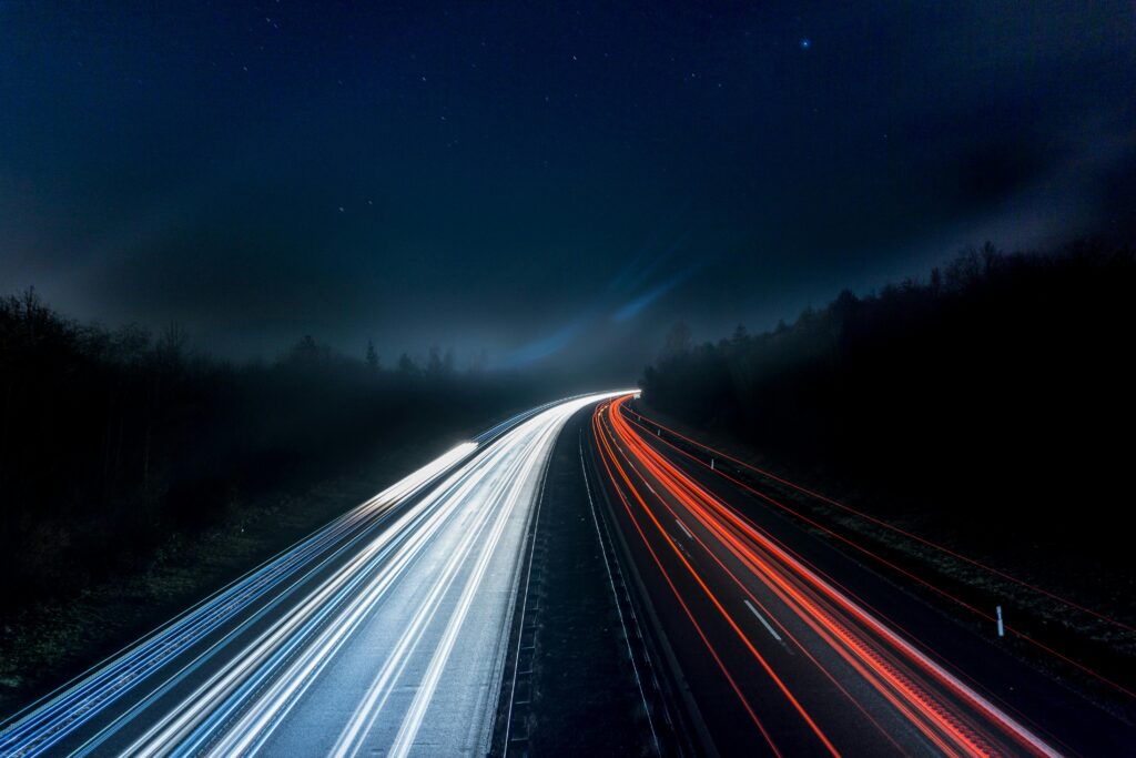 A motorway with blurred headlights to show the moving car. It is dark and surrounded by trees. 