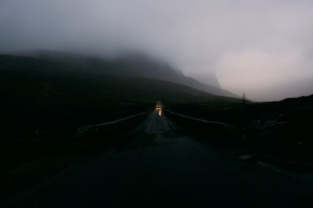 A dull road in a mountainous areas with a singular car driving down it with its headlights on. Mountains can be seen in the background, surrounded by fog and clouds. 